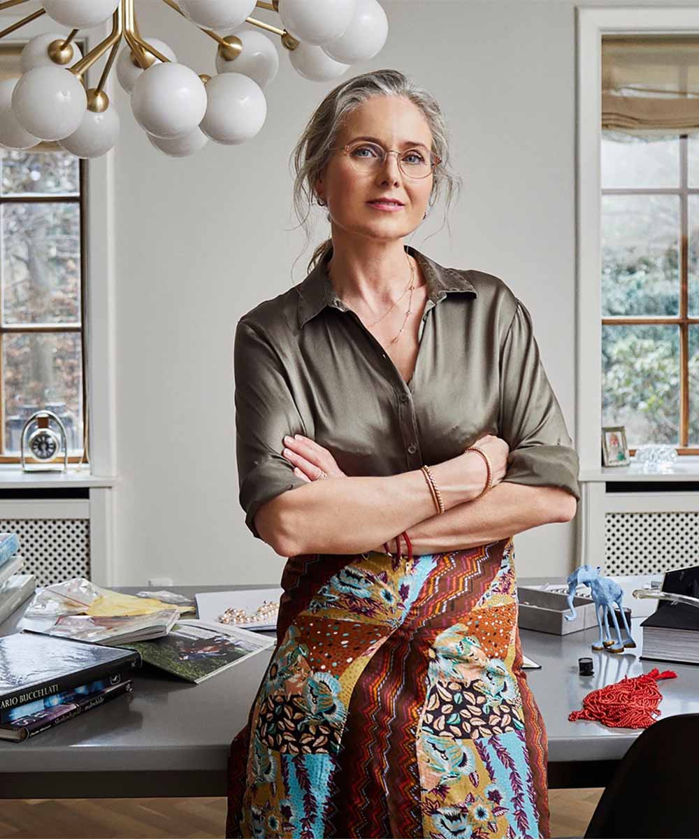 A woman wearing Bolou jewelry stands with her arms crossed in a bright room.